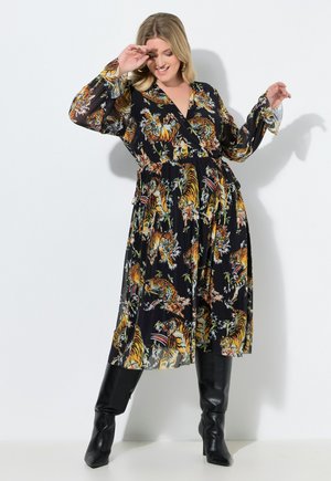 Vrouw in een zwarte midi-jurk met kleurrijke tijgerprint en lange mouwen, draagt zwarte kniehoge laarzen, poseert tegen een witte achtergrond.