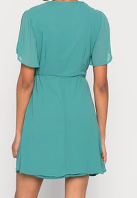 Robe courte teal en tissu léger, dotée d'une taille cintrée, de manches courtes bouffantes et d'une jupe évasée. Texture lisse sans motifs.