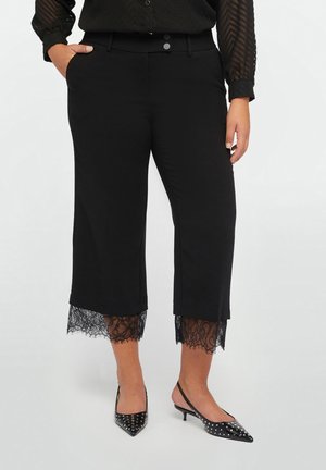Pantalon noir court avec bordure en dentelle aux ourlets, associé à des talons noirs cloutés à bride arrière et un haut noir transparent à manches longues.