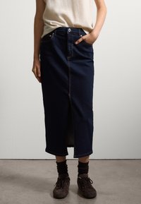 Falda de mezclilla en azul oscuro, hasta la rodilla y con una abertura frontal. Cuenta con un cierre de botón y cremallera, diseño de cinco bolsillos y costuras en contraste.