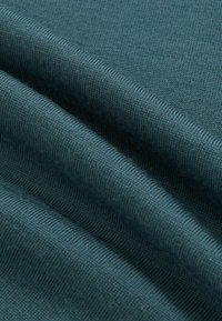 Detailní záběr na tyrkysovou pletenou tkaninu s viditelnou texturou a jemnými záhyby.