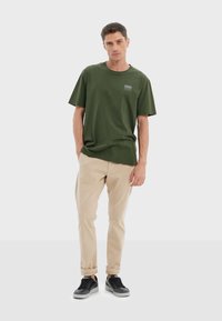Pantalones caqui, camiseta verde de algodón con un pequeño logo en la parte delantera y zapatillas negras. Diseño simple, casual y de corte relajado.
