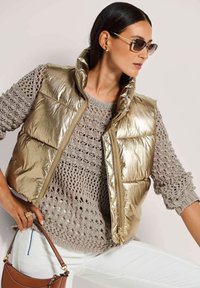 Metallisch goldene Steppweste mit hohem Kragen, tailliertem Schnitt und Reißverschluss. Kombiniert mit einem hellgrauen, dick gestrickten Pullover und weißen Hosen.