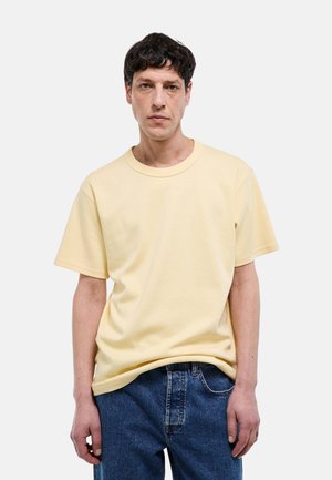 Homme portant un t-shirt à manches courtes jaune pâle et un jean bleu, debout devant un fond uni gris clair.