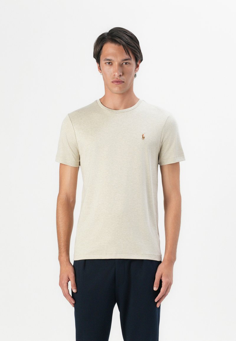 Joven de pie, vistiendo una camiseta de manga corta beige claro con un pequeño logo bordado en el pecho y pantalones oscuros.