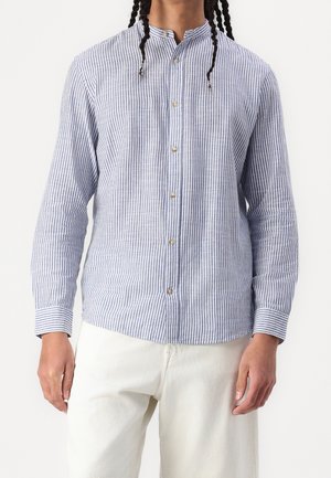 Homme portant une chemise à manches longues boutonnée, rayée verticale bleue et blanche, avec un pantalon blanc, debout devant un fond uni.