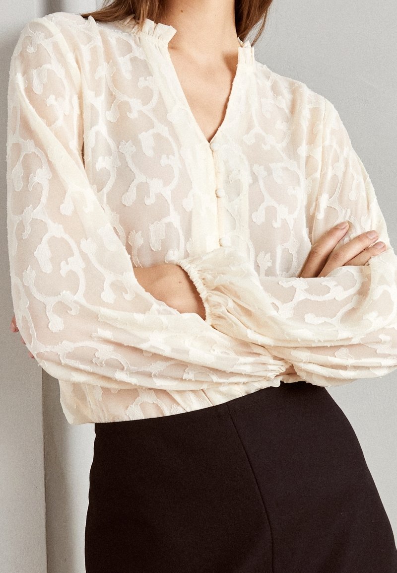 Blouse en crème transparente avec un motif floral texturé, fermeture à boutons sur le devant, col montant et poignets froncés. Associée à un bas noir.