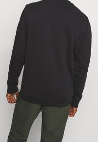 Hombre con una camiseta negra de manga larga simple y pantalones verde oscuro, visto desde atrás sobre un fondo neutro.