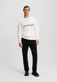 Balta megzta suknelė su apvaliu kaklu, su juodu š embroidered "Karl Lagerfeld" logotipu priekyje, derinama su juodomis kelnėmis ir bateliais.