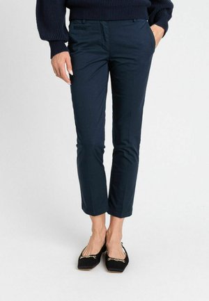 Donna che indossa pantaloni aderenti alla caviglia di un blu navy scuro, ballerine nere con dettagli dorati e un maglione a maniche lunghe blu navy scuro, in piedi con le mani nelle tasche.