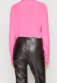 Camicia a maniche lunghe tagliata in tessuto rosa vivace, abbinata a pantaloni a vita alta in finta pelle nera con cuciture laterali e una texture lucida.