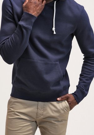 Mann trägt marineblauen Kapuzenpullover mit weißer Kordel und beige Hose, eine Hand berührt das Gesicht, die andere in der Tasche, vor einfarbigem Hintergrund.