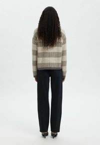 Personne avec de longs cheveux bouclés portant un pull rayé beige et gris, un jeans noir à jambes larges et des chaussures à talons noirs, se tenant de dos.