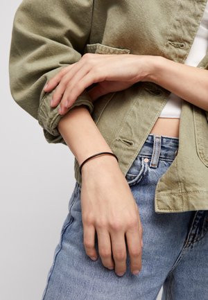 Bracelet en métal brun fin porté au poignet, posé sur une veste en denim verte et un jean bleu clair. La main a un teint de peau naturel.