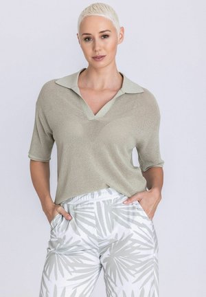 Vrouw met kort blond haar, gekleed in een licht beige gebreide top met kraag en witte broek met grijze bladpatronen, handen in de zakken.