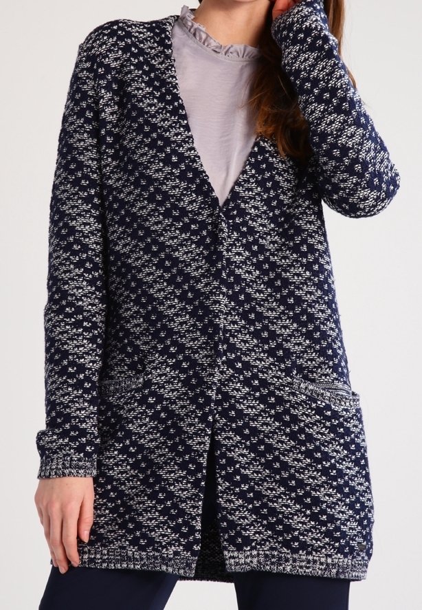 Femme portant un cardigan tricoté à motif bleu marine et blanc sur un haut gris clair à col à volants, la main reposant près du visage.