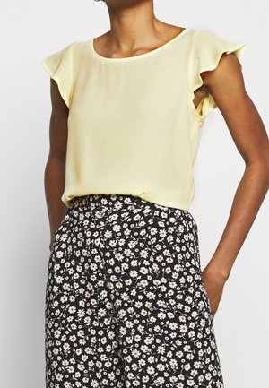Jasnożółta bluzka z krótkimi, falbaniastymi rękawami, noszona z czarnymi spodniami w kwiaty, ozdobionymi białymi i pomarańczowymi stokrotkami.