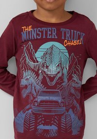 Bordeauxrotes Langarmshirt mit blauen und orangenen Dinosauriermotiven, einem Monstertruck und dem Text "DIE MONSTERTRUCK-VERFOLGUNG!" in fetter Schrift.