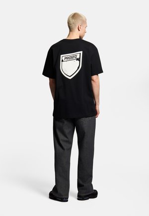 Mężczyzna z krótkimi blond włosami, ubrany w luźną czarną koszulkę z białym logo w kształcie tarczy „PROSTO” na plecach, ciemne dżinsy i czarne buty, stojący.