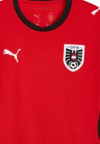 Κόκκινη αθλητική φανέλα με μαύρες πινελιές, με λευκό λογότυπο Puma στα αριστερά και ένα κοντόσχημο έμβλημα με σχέδιο ποδοσφαιρικής μπάλας.