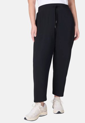 Personne portant un pantalon noir ample avec cordon et des baskets blanches, vue de la taille aux pieds sur un fond uni.