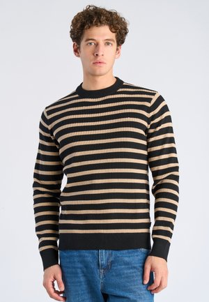 Jonge man met krullend haar, gekleed in een zwart-beige trui met horizontale strepen en een blauwe spijkerbroek, staand tegen een effen lichte achtergrond.