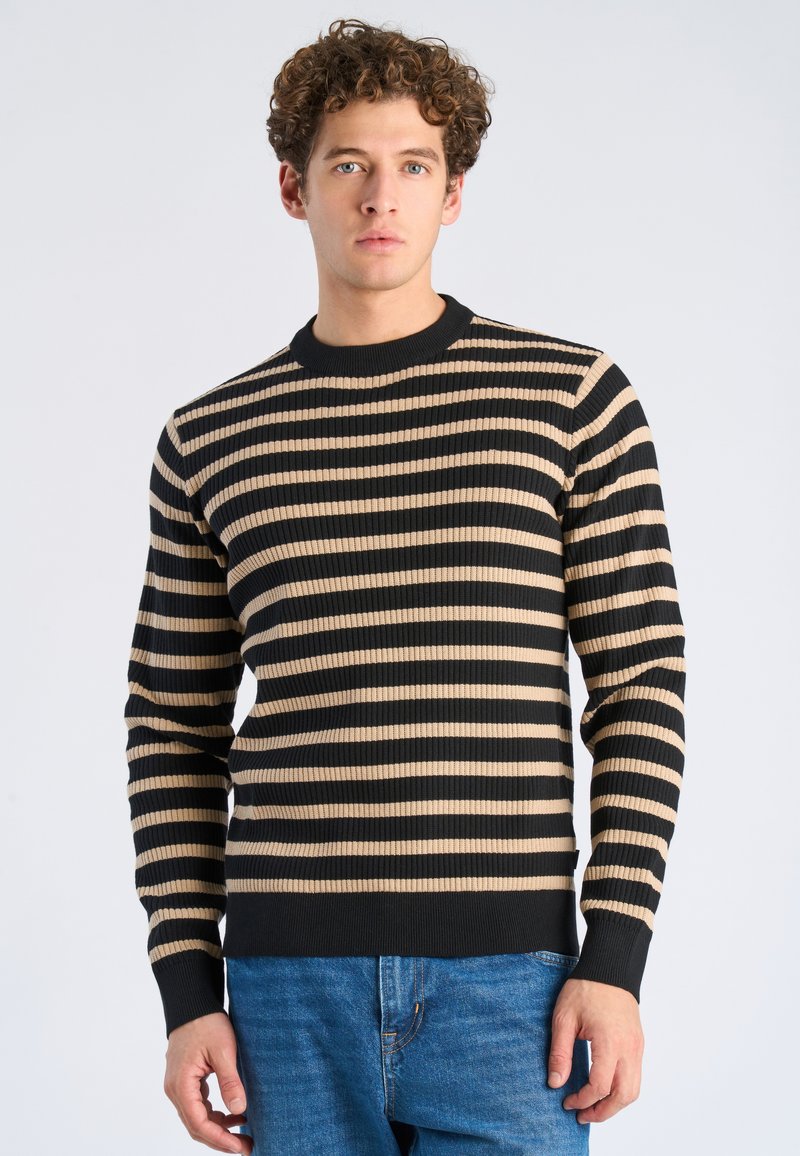 Młody mężczyzna z kręconymi włosami, ubrany w czarno-beżowy sweter w poziome paski i niebieskie dżinsy, stojący na tle jasnej, jednolitej ściany.