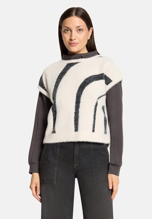 Woman with straight dark hair wears a white sleeveless sweater with black abstract stripes over a long-sleeve dark gray shirt and black jeans.