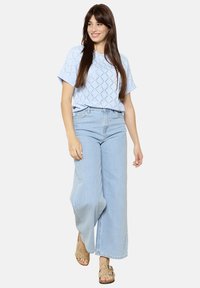 Svetlomodrý pletený top s geometrickým vzorom, skombinovaný so svetlomodrými širokými džínami. Outfit dopĺňajú béžové sandále so dekoratívnymi cvokmi.