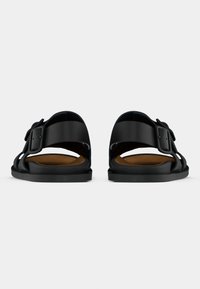 Μαύρα σανδάλια με φαρδύ λουράκι και ρυθμιζόμενο αγκράφα, επίπεδη σόλα, ανοιχτή πλάτη και ομαλή υφή. Στο εσωτερικό διαθέτει καφέ επένδυση.