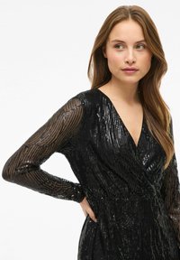 Black wrap dress with long, sheer sleeves, adorned with sequins in a vertical pattern, featuring a deep V-neckline and gathered waist.