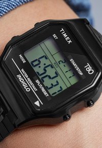 Orologio digitale nero con cassa esagonale, dotato di display retroilluminato verde, controlli etichettati per modalità, impostazione e avvio/arresto, indossato a un polso.