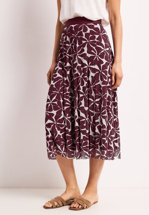 Kvinde iført en plisseret midi-nederdel med stort mørkelilla blomsterprint og tan farvede flade slidesandaler, stående mod en ensfarvet lys baggrund.