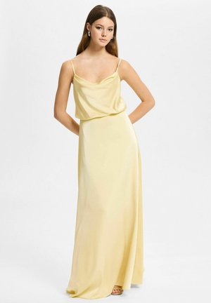 Młoda kobieta o długich brązowych włosach, ubrana w bladożółtą satynową sukienkę na ramiączkach i kolczyki koła, stojąca z rękami za plecami.