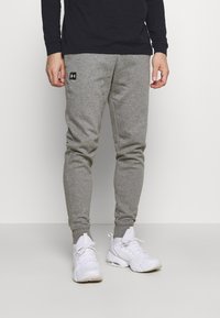 Graue sportliche Jogginghosen mit elastischen Bündchen und einem schwarzen Logo-Patch. Kombiniert mit weißen Sneakers und einem schwarzen Langarmshirt.