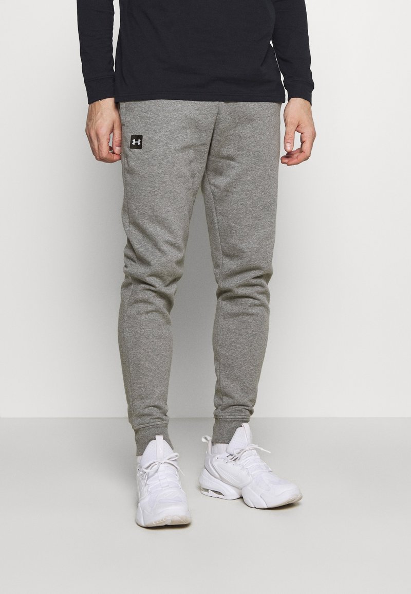 Graue sportliche Jogginghosen mit elastischen Bündchen und einem schwarzen Logo-Patch. Kombiniert mit weißen Sneakers und einem schwarzen Langarmshirt.