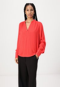 Blusa rossa con scollatura a V e sottili righe verticali. Presenta maniche lunghe con polsini arricciati. Abbinata a pantaloni neri. Texture liscia.