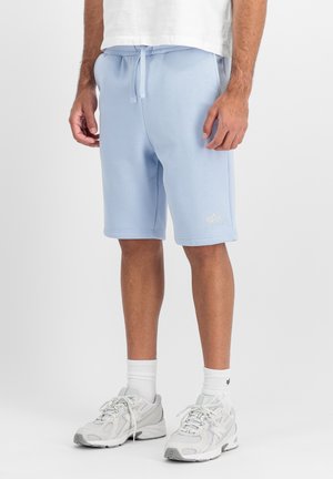 Mann trägt hellblaue Shorts mit Kordelzug, weiße Knöchelsocken und weiße Sportschuhe, steht vor einem einfarbigen Hintergrund.