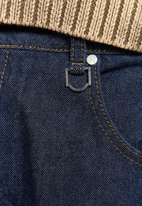 Jeans in denim blu scuro con cuciture visibili, caratterizzati da un bottone in metallo e un design unico della pattina sopra la tasca anteriore. Maglione in maglia testurizzata sopra.