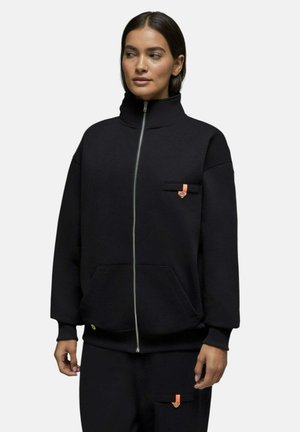 Femme portant une veste noire zippée et un pantalon assorti avec des détails orange sur les poches, debout devant un fond uni.