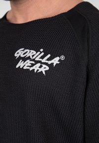 Zwarte textuurtrui met een geribbeld patroon. Dikke witte "GORILLA WEAR" logo op de borst, met platte naden en een ronde hals.