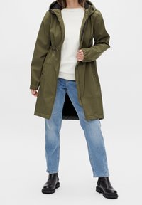 Manteau imperméable vert olive avec capuche, taille à cordon, manches longues et fermeture éclair devant, assorti à un jean bleu clair et des bottes noires.