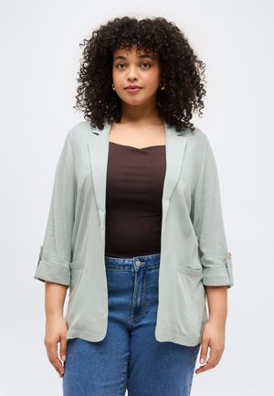 Kvinne med krøllete hår som har på seg gullfargede hoops-øredobber, en lys grå åpen blazer, mørkebrun topp og blå jeans, vendt fremover mot en hvit bakgrunn.