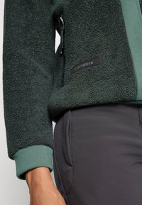 Close-up of a person wearing a dark green textured fleece jacket and dark gray pants, showing zipper and pocket details.