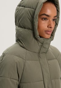 Töltött olajzöld dzseki magas gallérral és nagy kapucnival. Steppelt textúrával és elől cipzáras záródással rendelkezik.