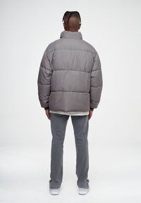 Doudoune grise avec un col haut, un design matelassé et des finitions élastiques, portée sur un pantalon gris clair et des baskets blanches.