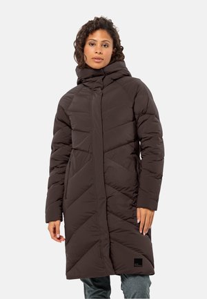 Langbruine pufferjas met een hoge kraag, een chevrond quiltpatroon en een logopatch aan de zoom. Het model heeft de handen in de zakken.