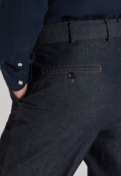 Dark blue denim pants with a textured finish, featuring contrasting orange stitching and a buttoned back pocket. A slim fit design is evident.