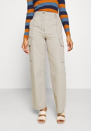 Femme portant un pantalon cargo beige à jambes larges, un haut à manches longues rayé multicolore et des sandales à talons ouvertes crème, debout sur un sol gris.