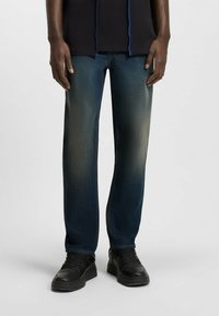 Jeans in denim blu con taglio dritto, caratterizzati da una finitura scolorita, cuciture evidenziate e texture usurata, abbinati a sneaker nere.